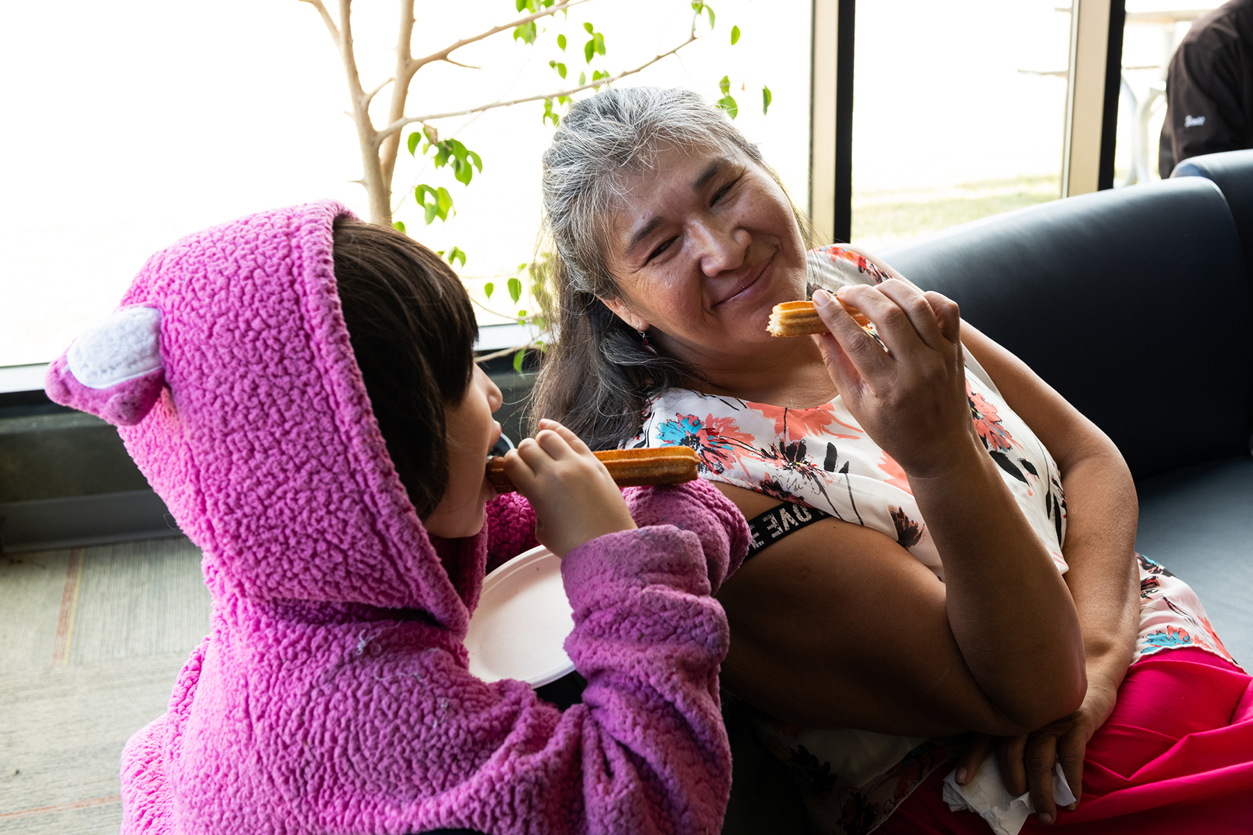 An Indigenous grandmother and granddaughter enjoying churros, sitting on a couch.