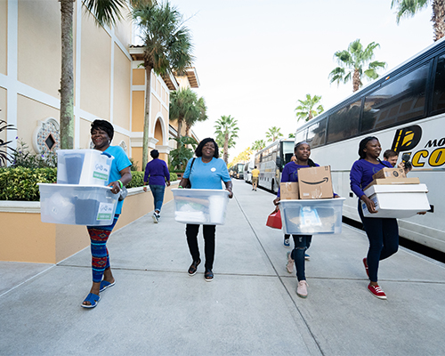 Four women carry supplies to be loaded onto a bus and donated during God in Shoes community service day. 