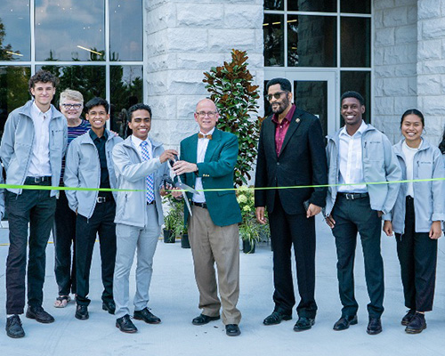 Eight people standing in front of a building. Two holding large scissors to cut a ribbon.