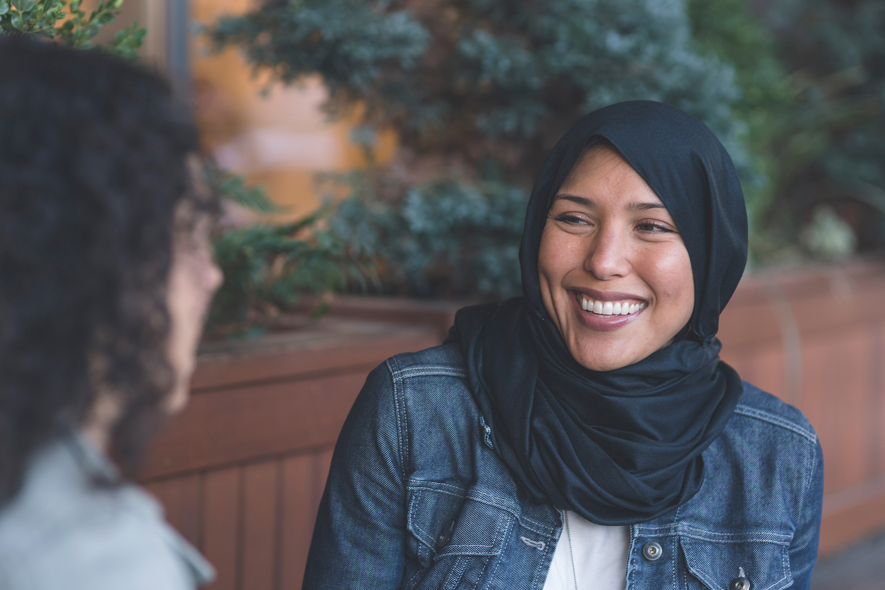 Muslim woman in a scarf smiling at another woman with curly hair whose face isn't shown. 