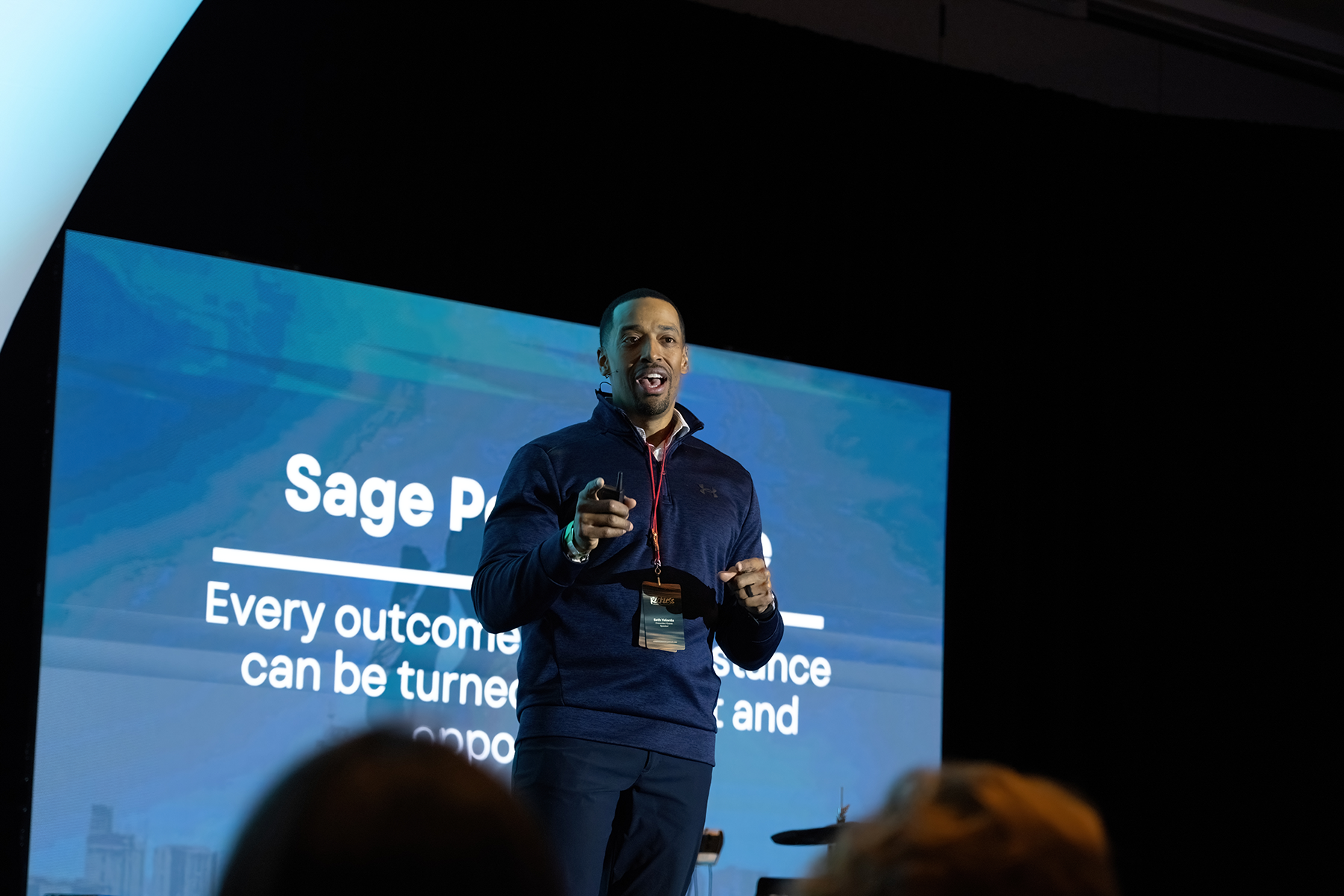 A black man stands in front of a large screen on a stage, speaking.