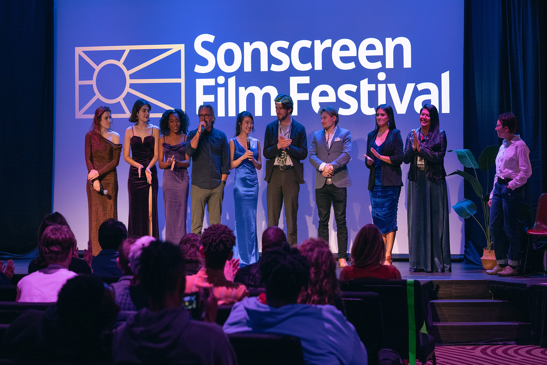 A diverse group of people stand on stage in front of an audience. The words "Sonscreen Film Festival" are behind them.