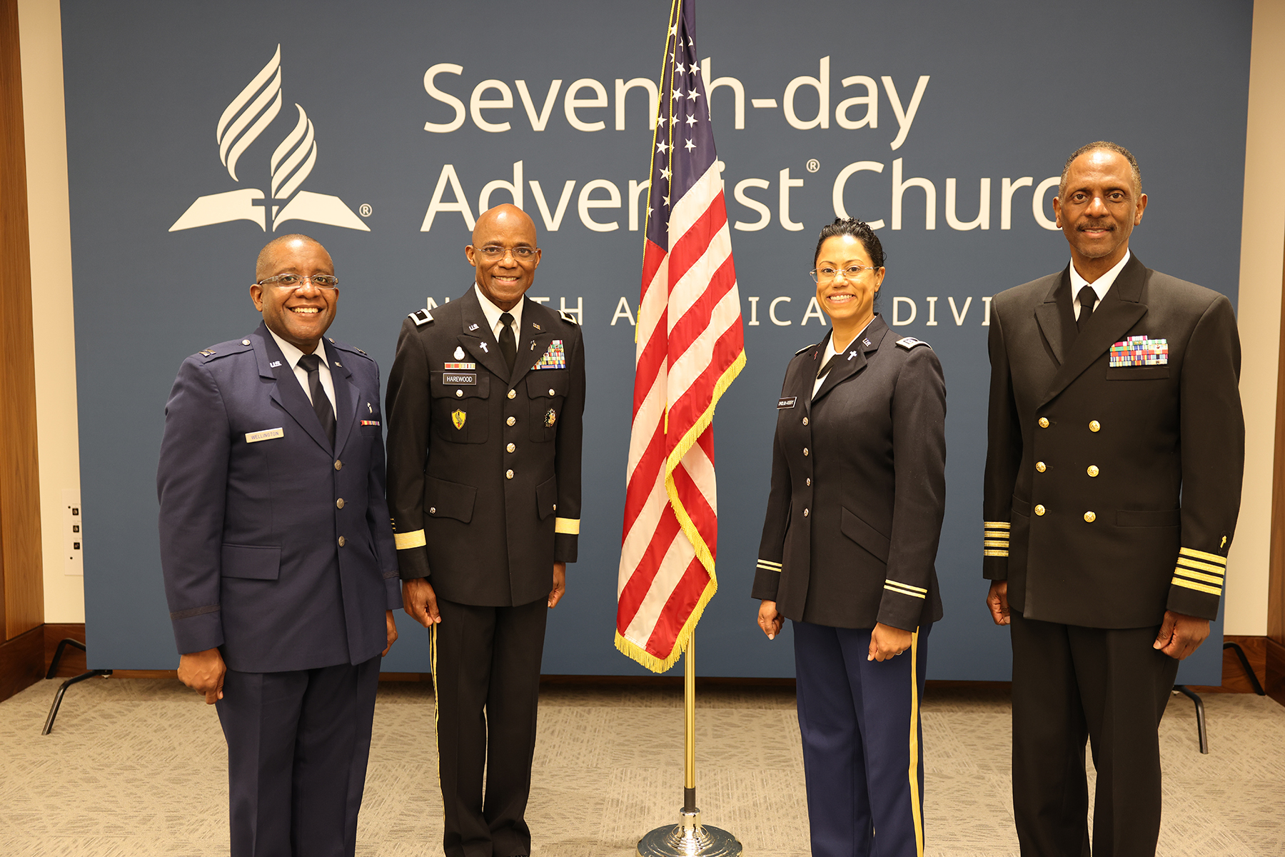 Four smiling individuals in military uniform with a U.S. flag in the middle of them; two black men, a woman with tanned skin, and another black man.