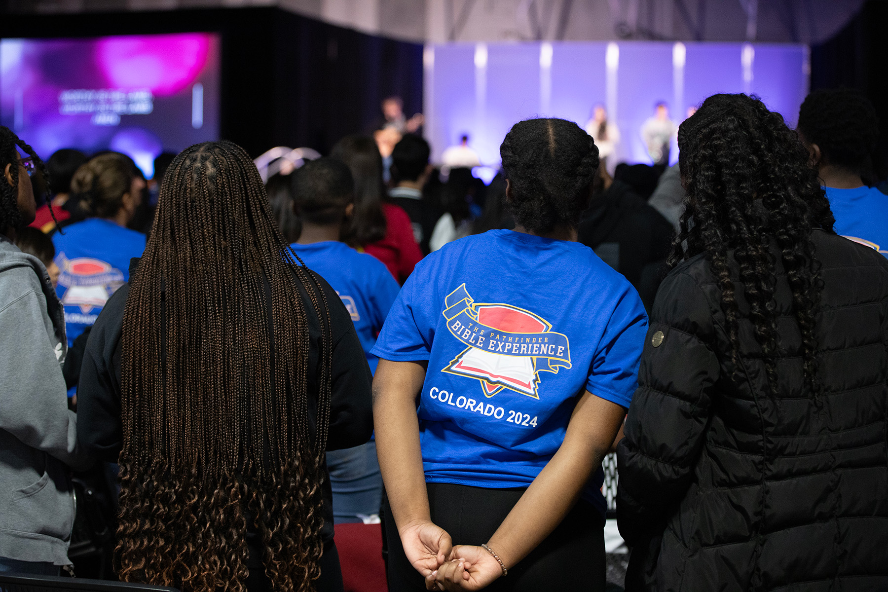 Shows the back of four black girls, standing up. In front of them is a blurred-out praise and worship team.