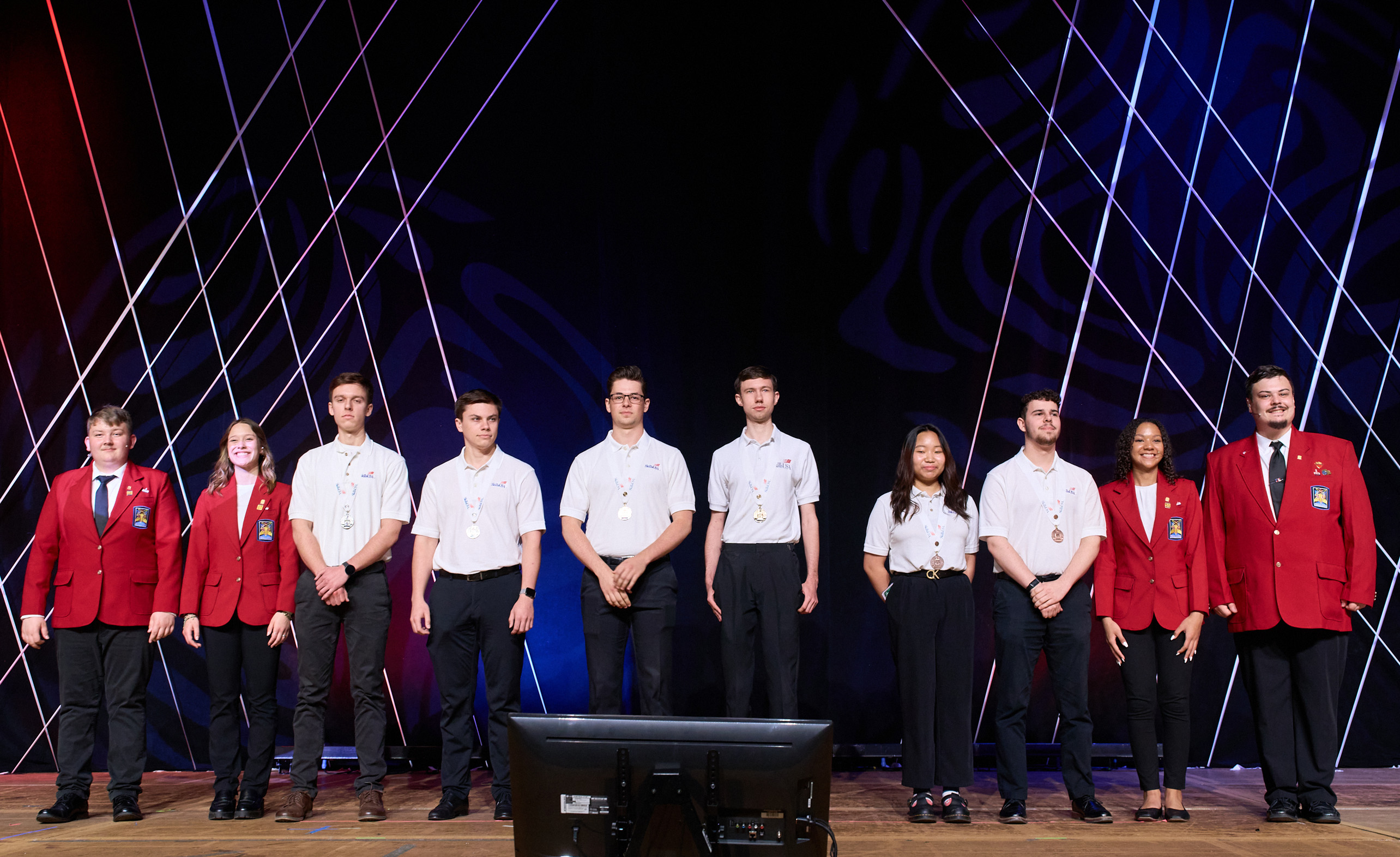 A diverse line-up of young men and women on a stage wearing white shirts, black pants and some with red jackets. 