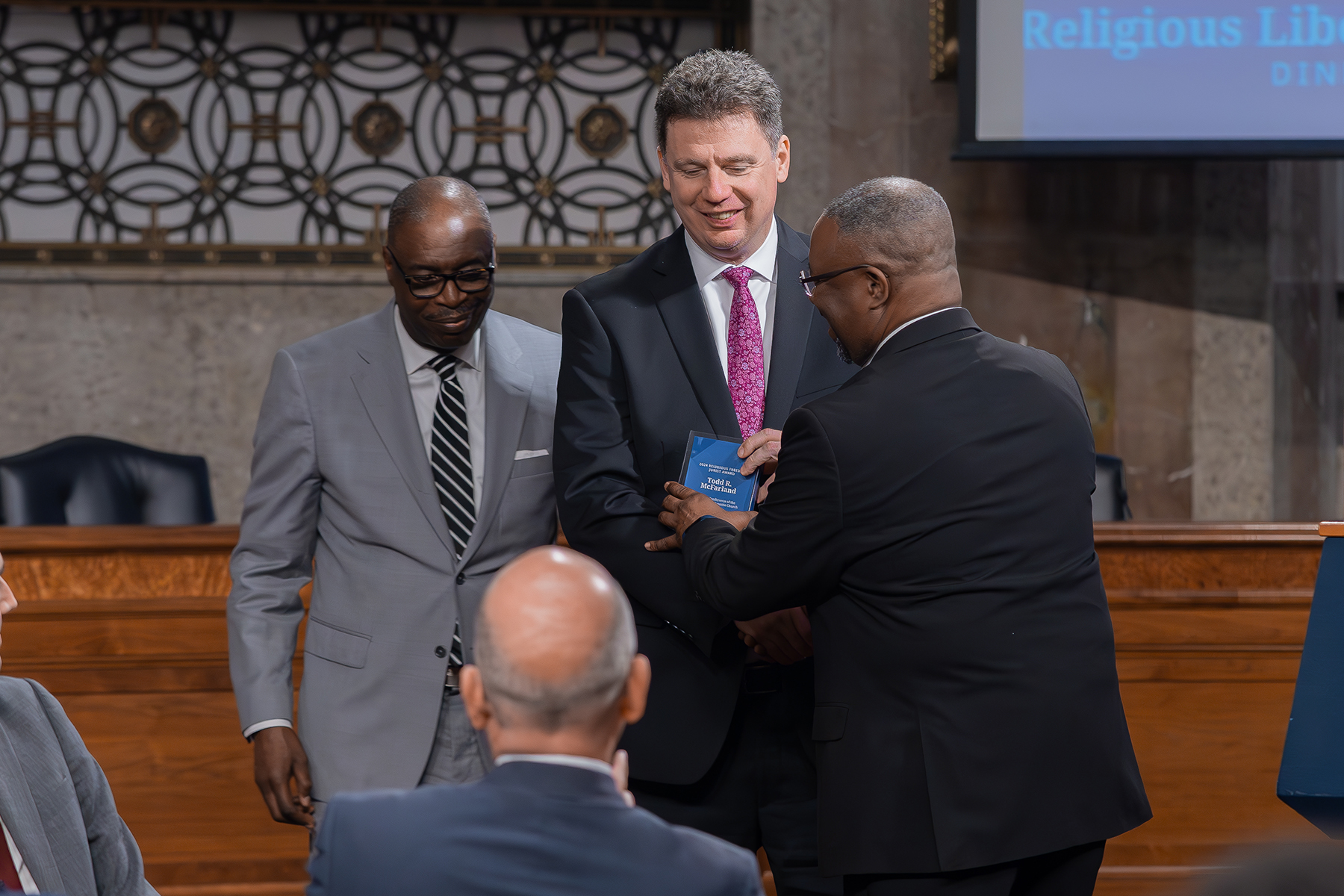 A white man receives an award from a black man as another black man looks on. 