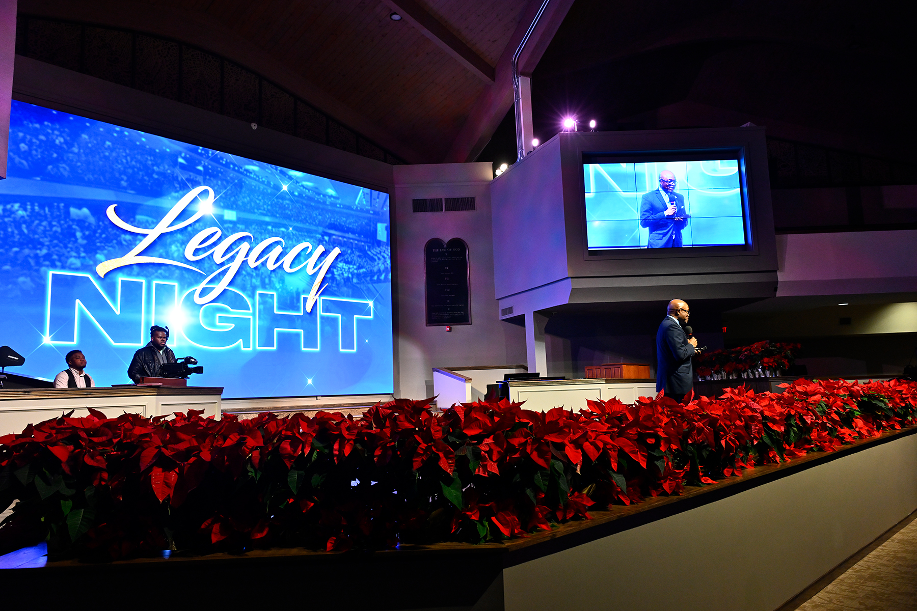 A black man on stage with his image on a small screen to the right of him and "Legacy Night" on a larger screen behind him.