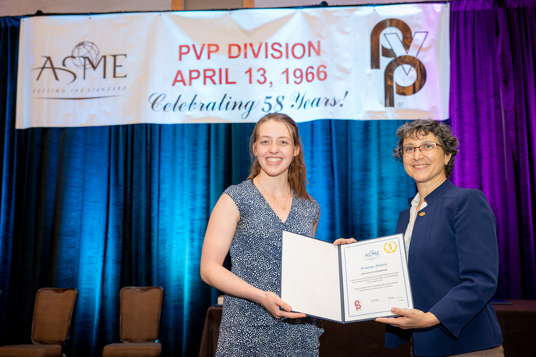 A young woman who has received an award (left) and the award presenter (right) both hold up the award.