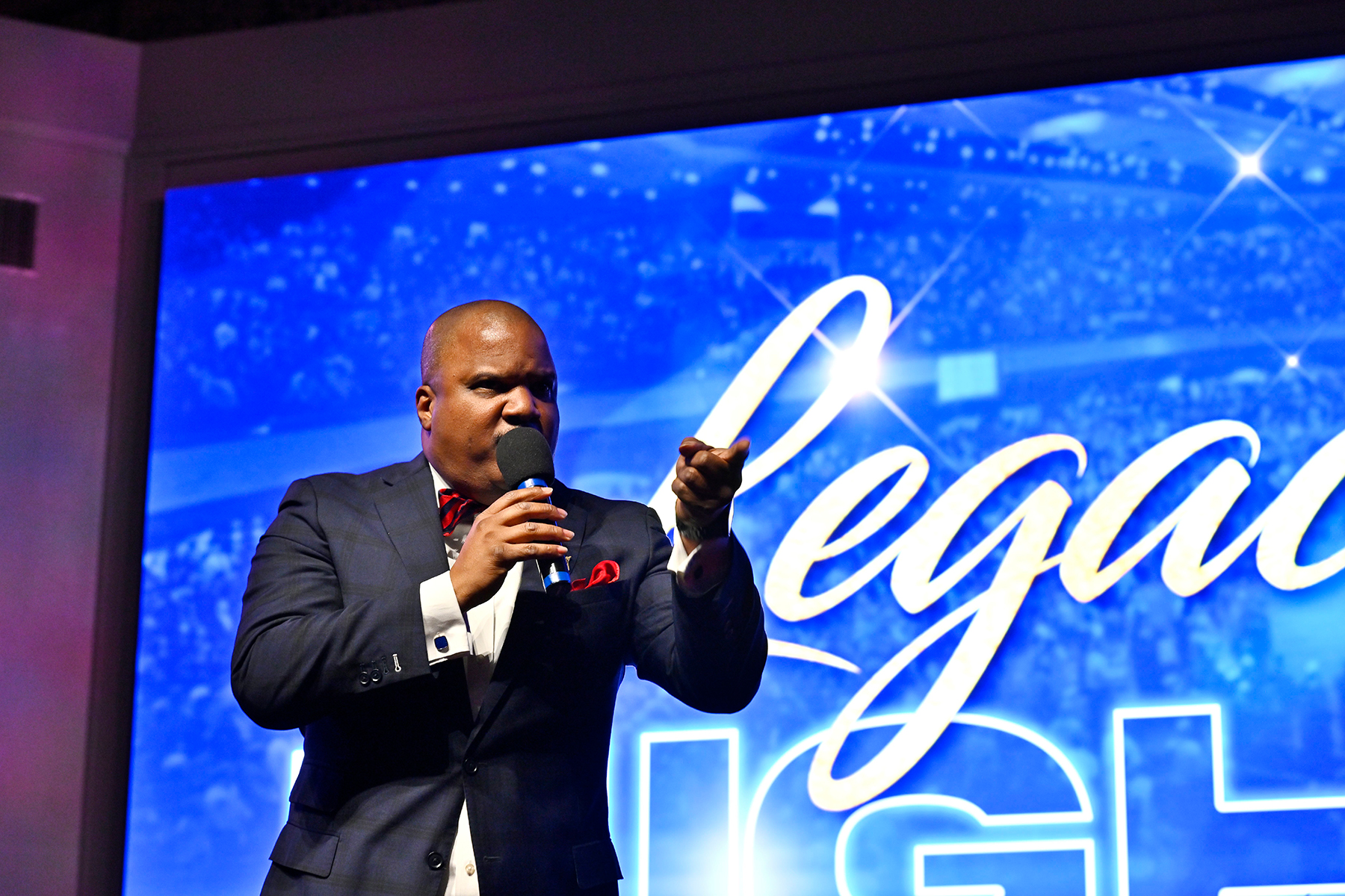 A black man preaches on stage in front of a screen reading "Legacy"