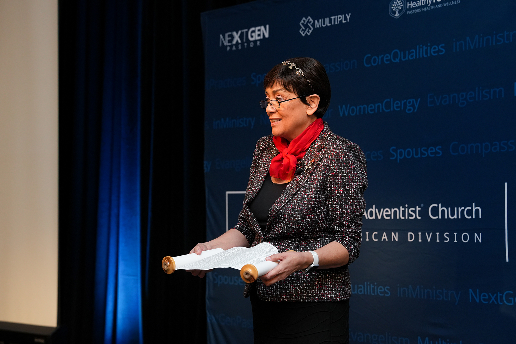 A Hispanic woman holding a scroll speaks at a conference.