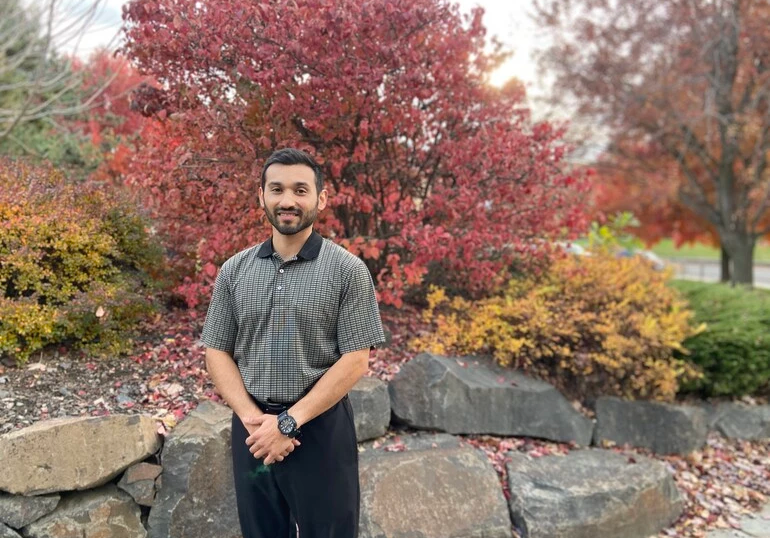 Young Hispanic man stands in front of a rock wall surrounding a garden.