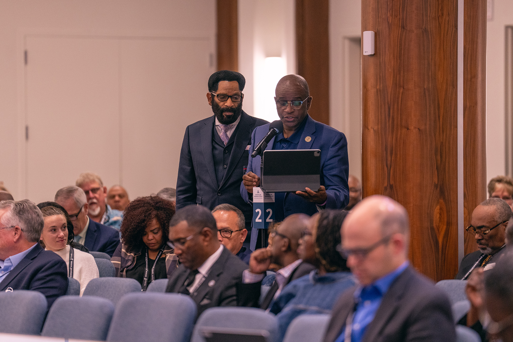 Two black men stand behind a microphone in an auditorium, one speaking. A diverse group of onlookers listen attentively. 