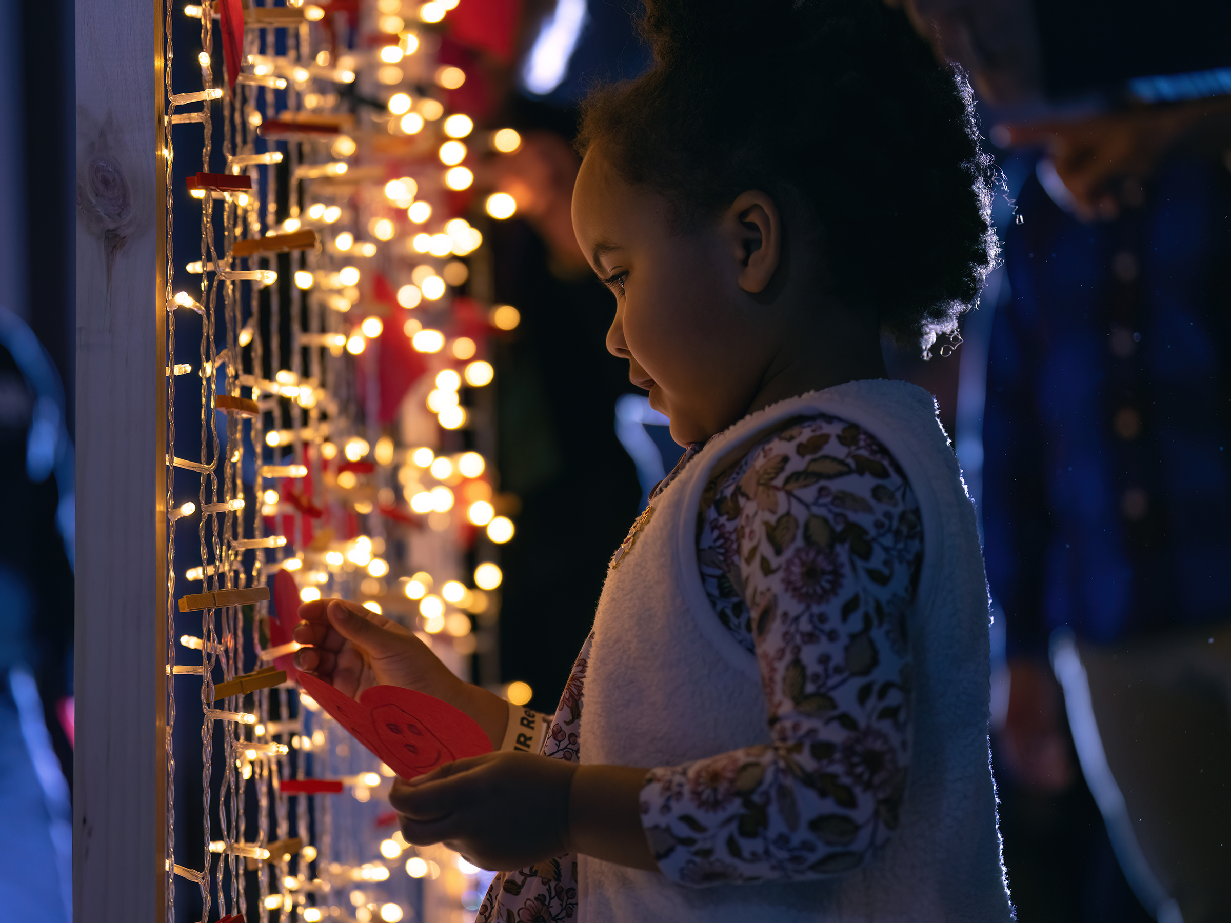 A little girl pins a heart with a picture on a wall of lights