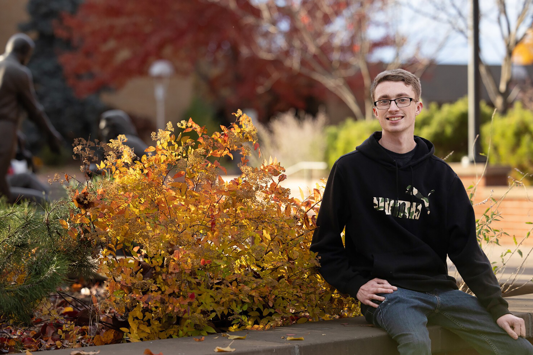 Bespectacled young man wearinga black hoodie sits in front of an orange bush. 