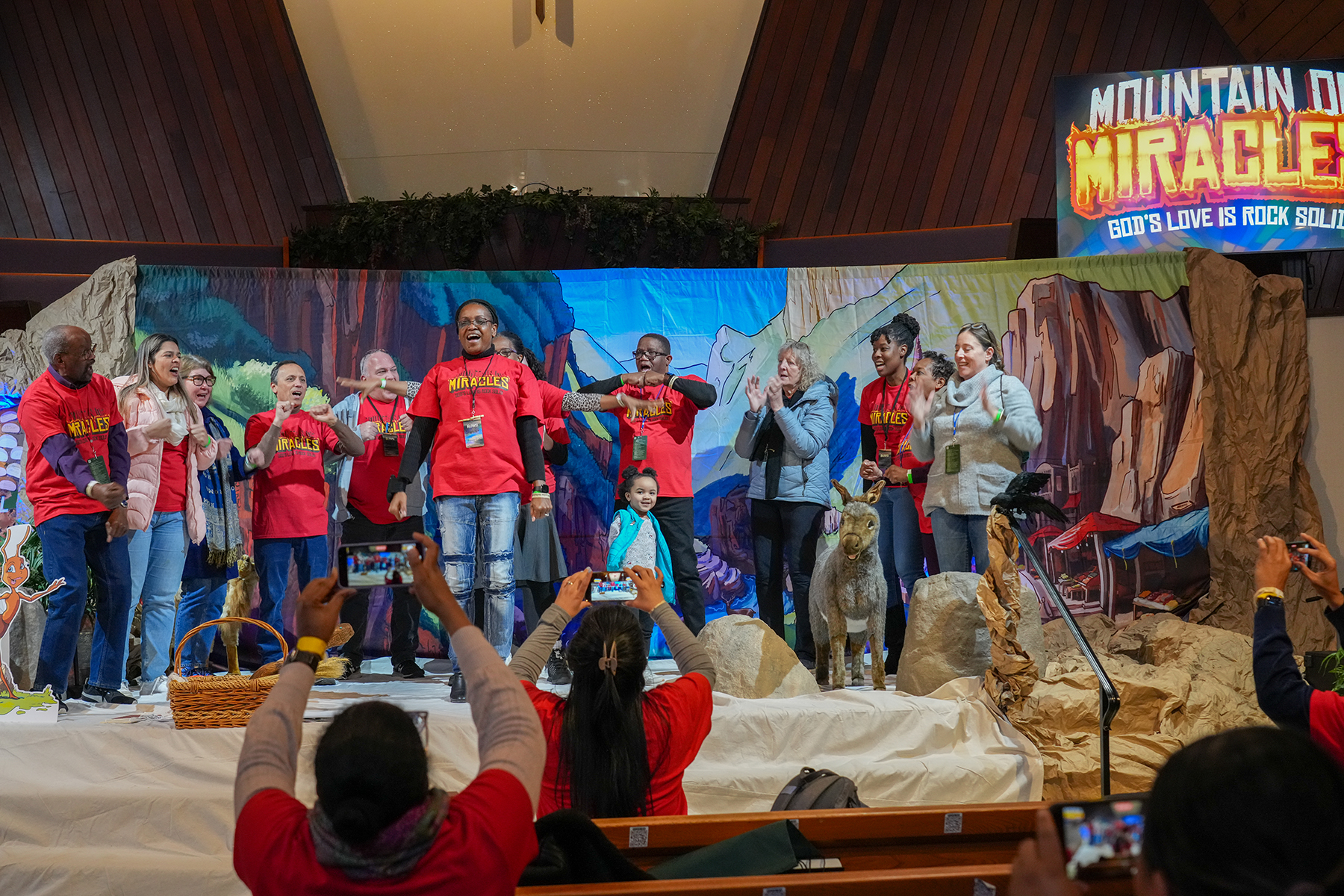 Diverse group on stage performing a cheer, with Mountain of Miracles logo behind them.