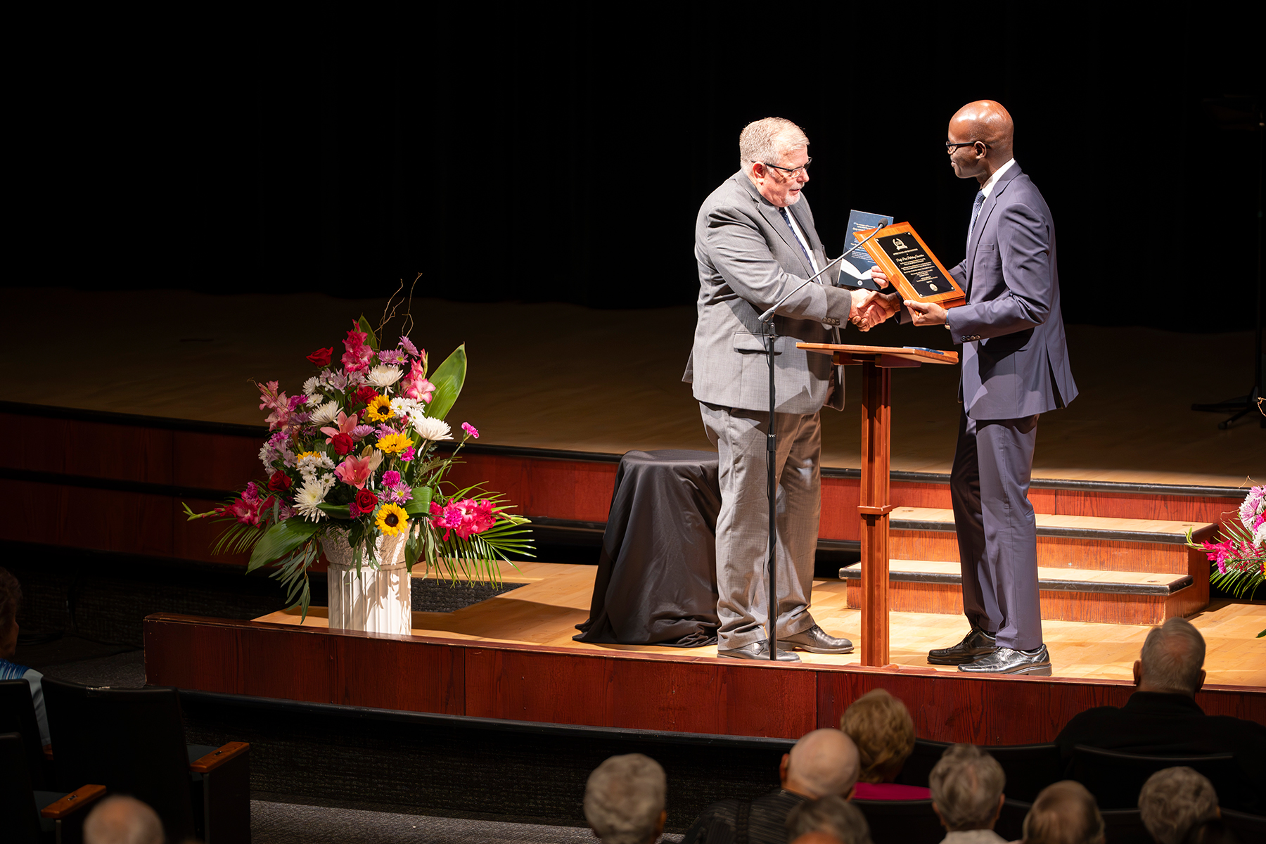 Two men of different races on a stage, and one man gives another a plaque as they shake hands