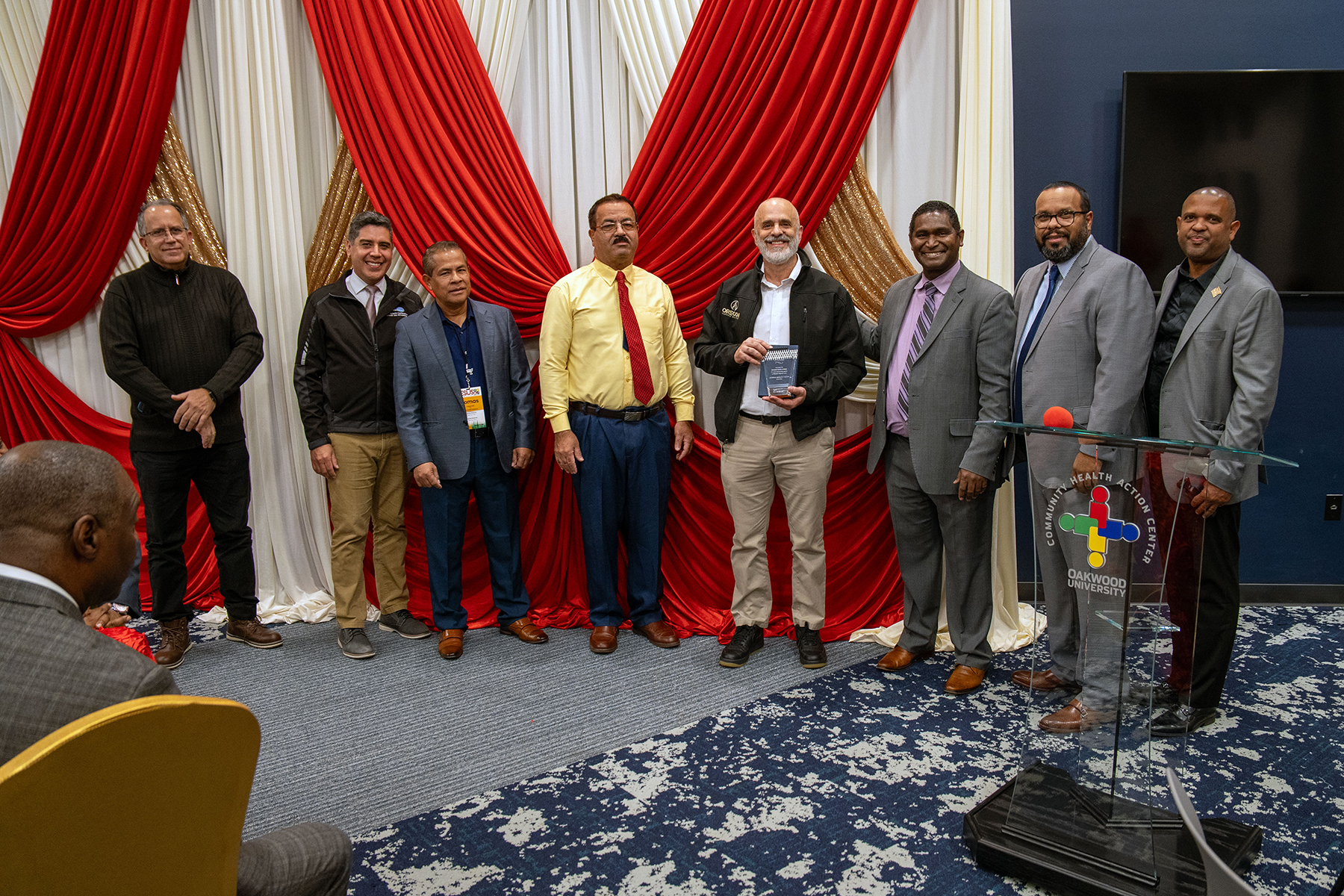 Eight men stand in front of a room with one holding a plaque. 