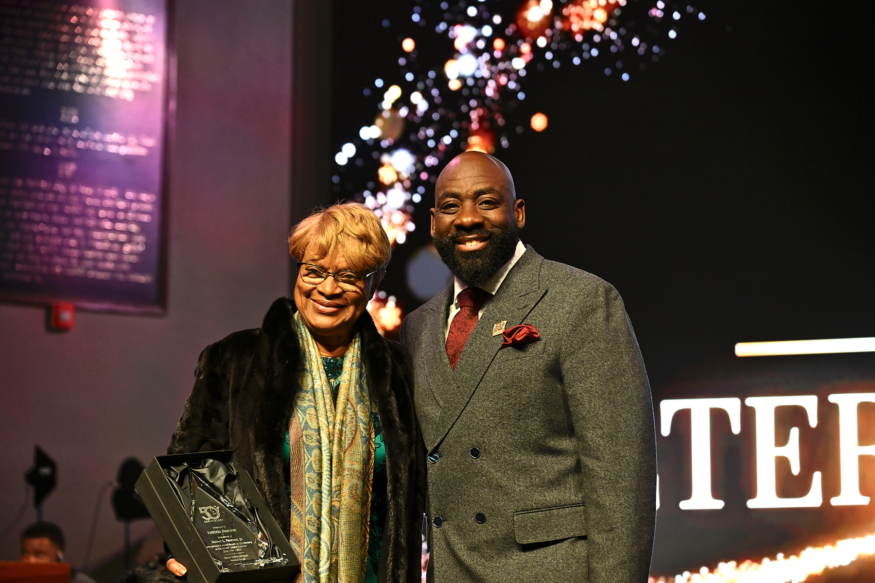 An older black woman and younger black man on stage, smiling. The woman holds a glass plaque. 