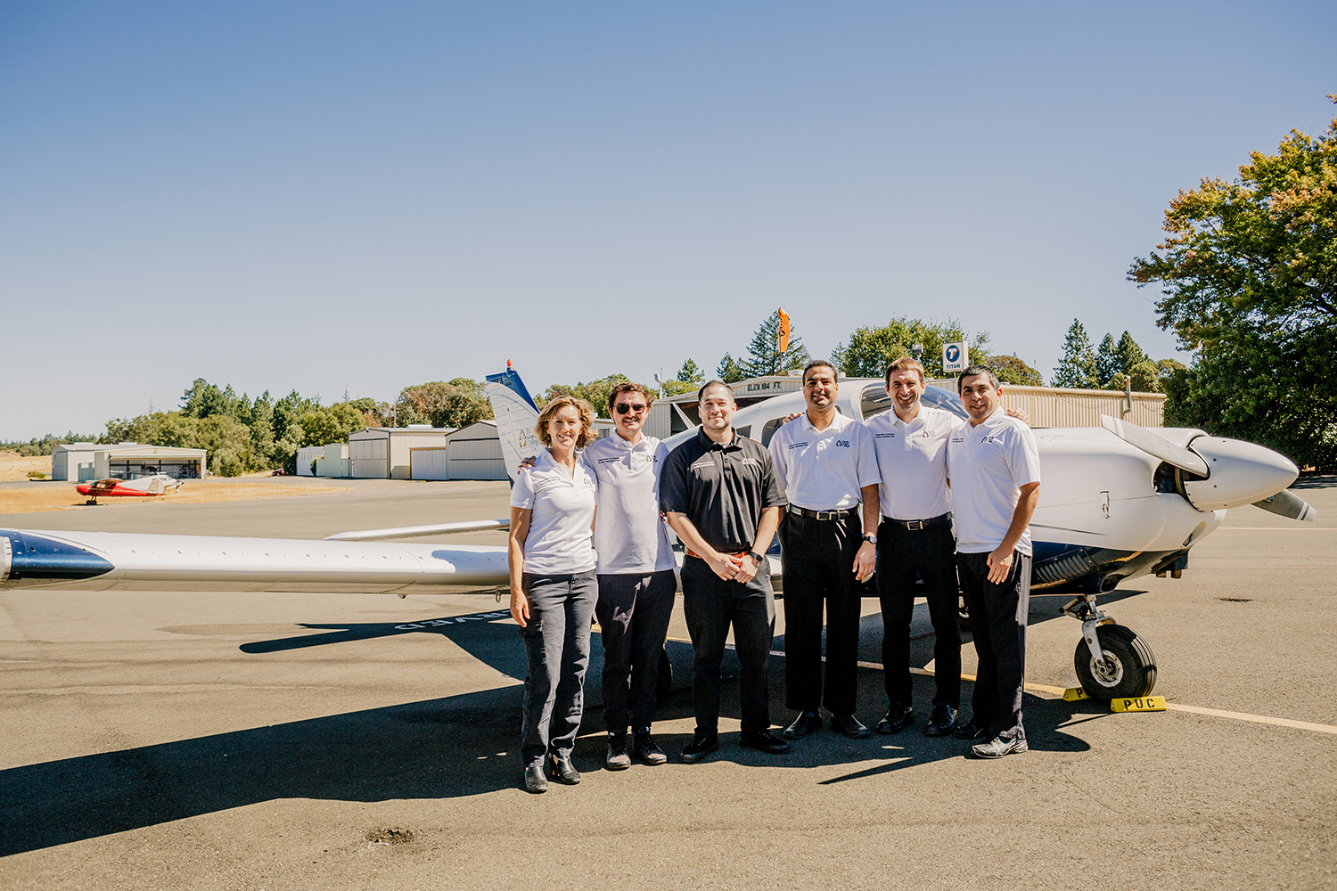 A diverse group of one woman and five men stand in  front of an airplane