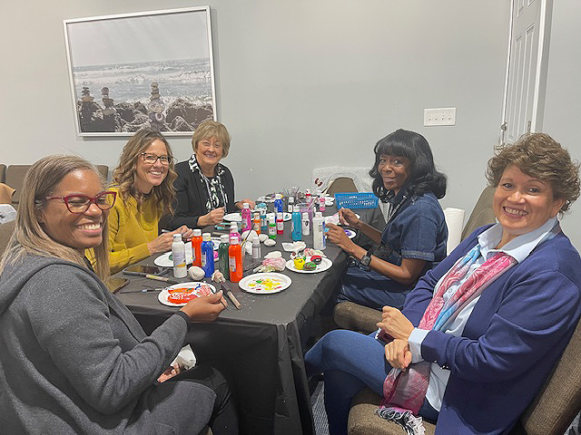 A diverse group of ladies sit at a table painting rocks. 