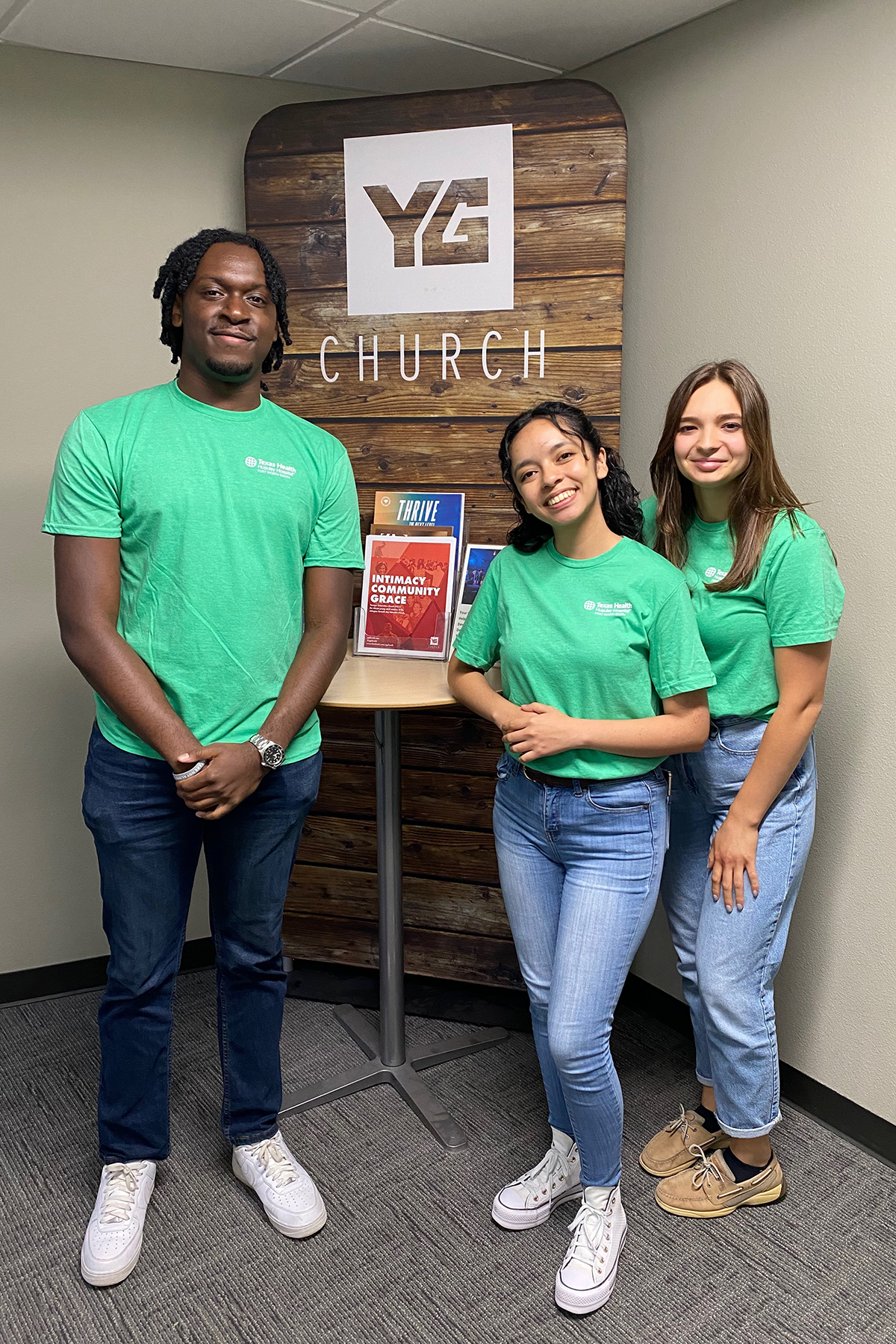 A young black man, Hispanic woman, and white woman standing in front of a wooden sign reading YG church wearing matching green AdventHealth